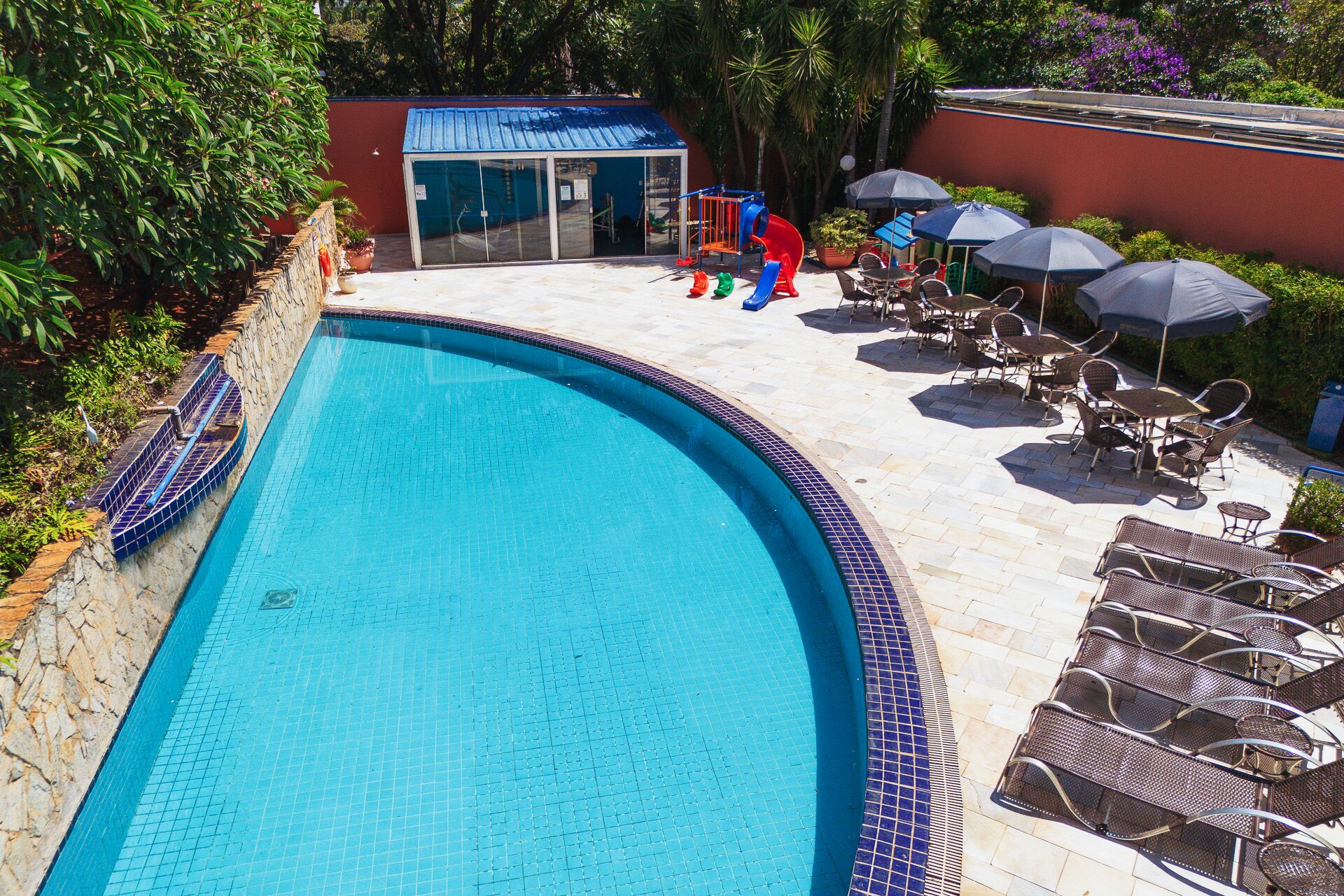 Una piscina con bordo piastrellato, circondata da lettini e ombrelloni, e una piccola area giochi con scivoli.