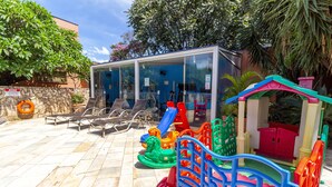 Zona de juegos infantiles con una construcción pequeña, toboganes y una zona de descanso junto a la piscina.