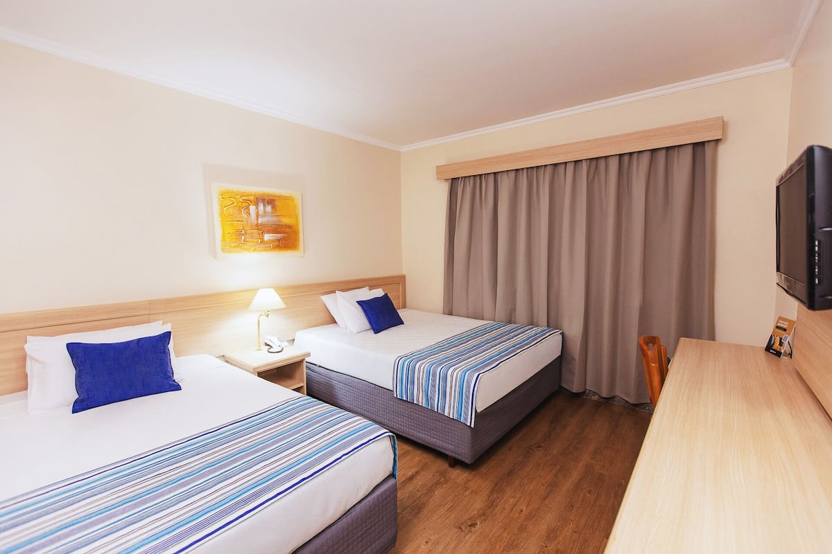 A hotel room with two beds, a wooden headboard, a bedside table with a lamp, a television, and a large window with curtains.