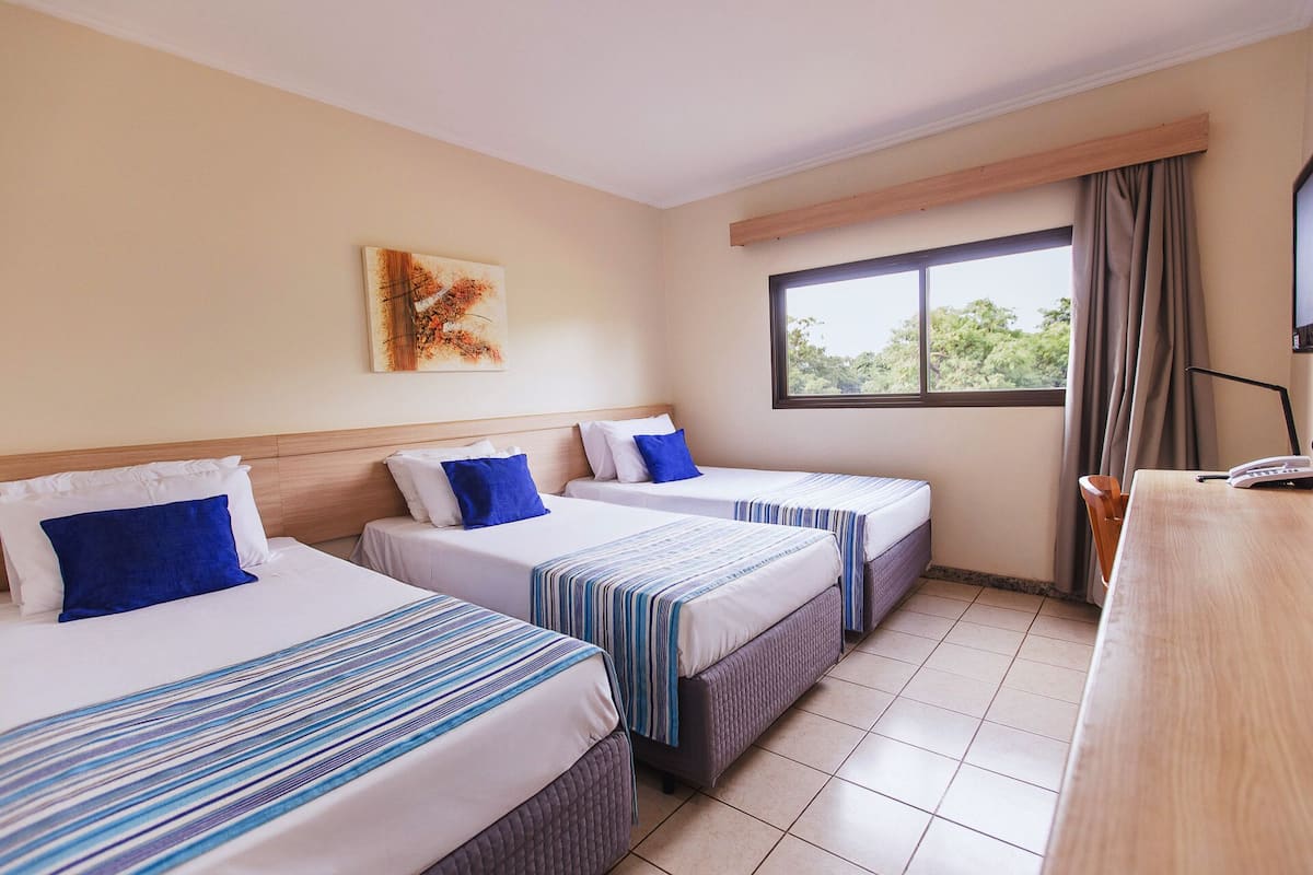 A hotel room with three beds, a window with a view, a painting on the wall, and a desk with a phone.