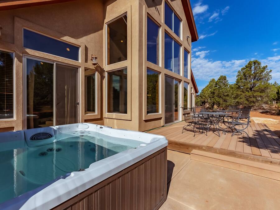 Un bain à remous sur une terrasse en bois, avec une table et des chaises d’extérieur, de grandes fenêtres et un ciel bleu dégagé.