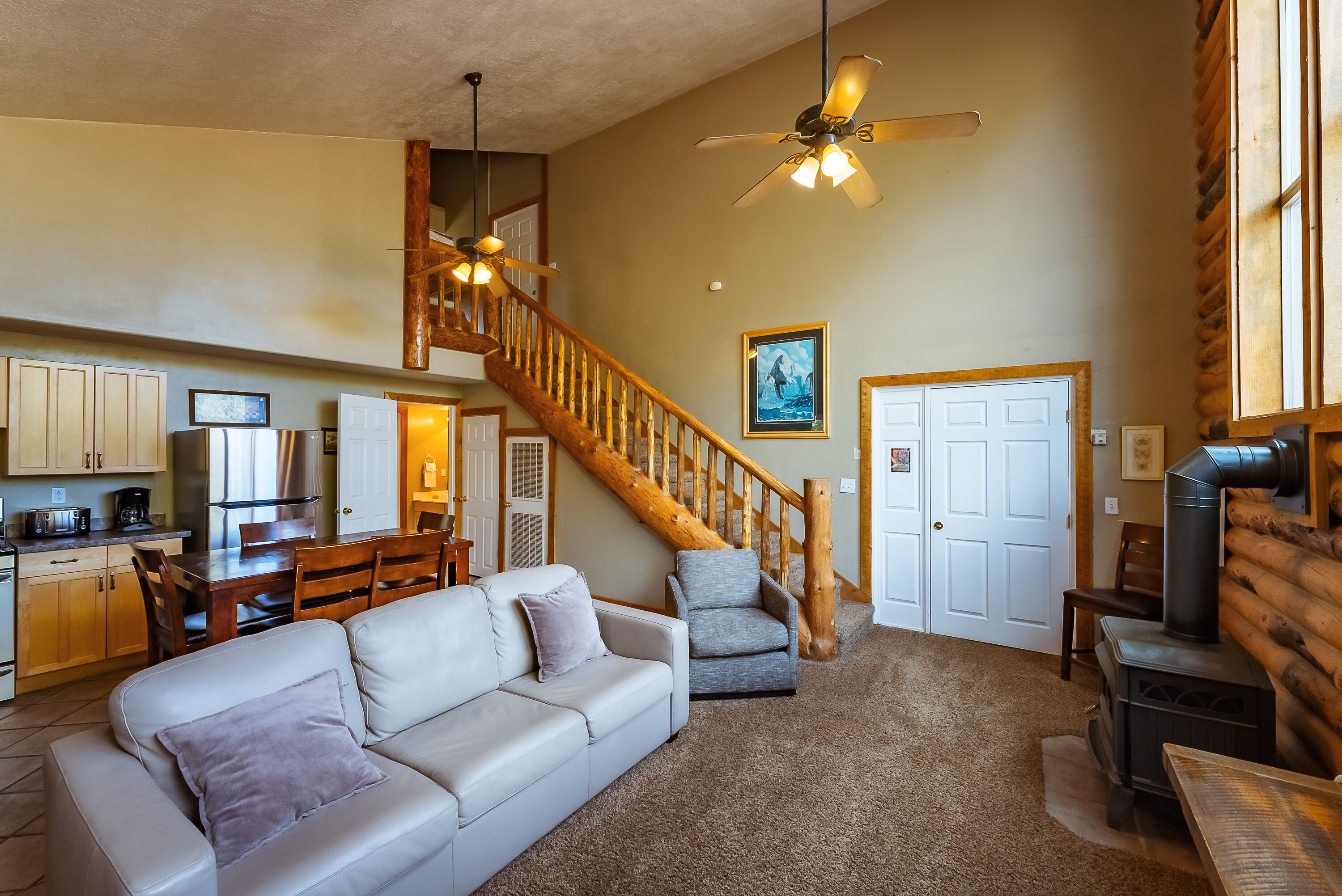 A spacious living area with a large white sofa, a grey armchair, a wooden staircase, and a wood-burning stove.