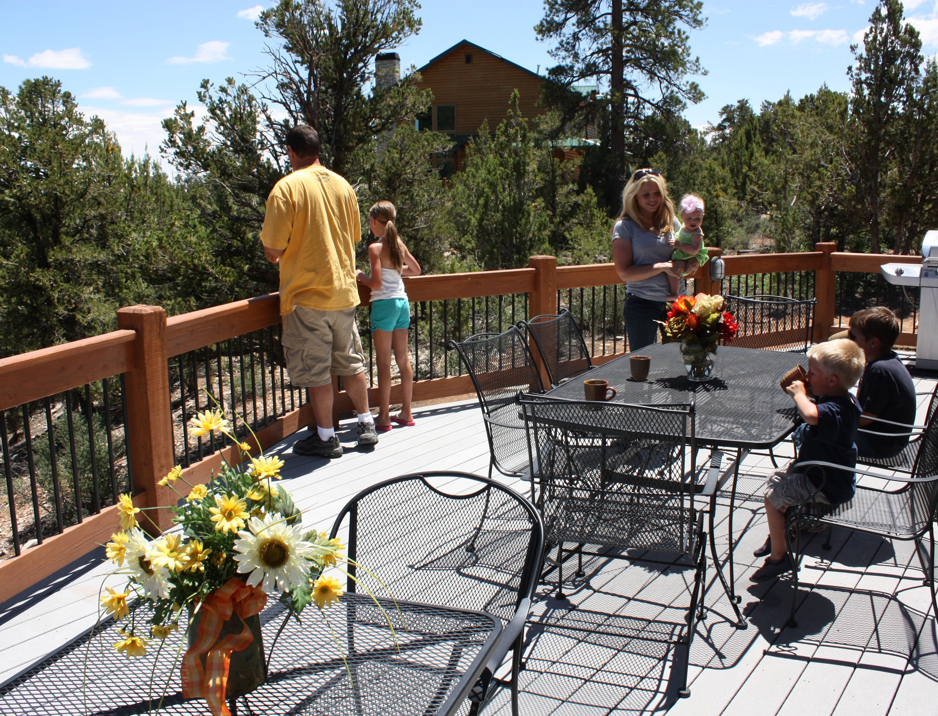 A deck with outdoor seating, a table with a vase of flowers, and a view of trees and a building in the background.
