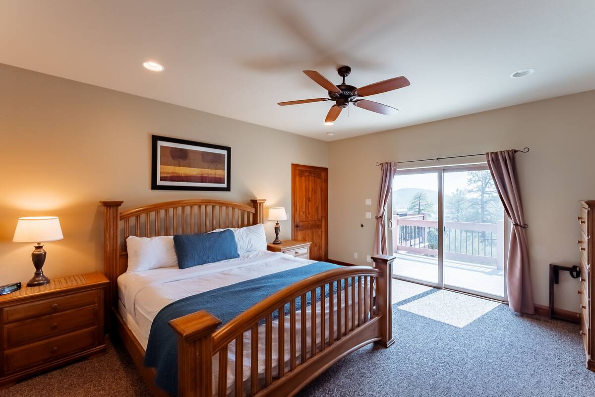 A bedroom with a wooden bed, a ceiling fan, a nightstand with a lamp, a window with curtains, and a door.