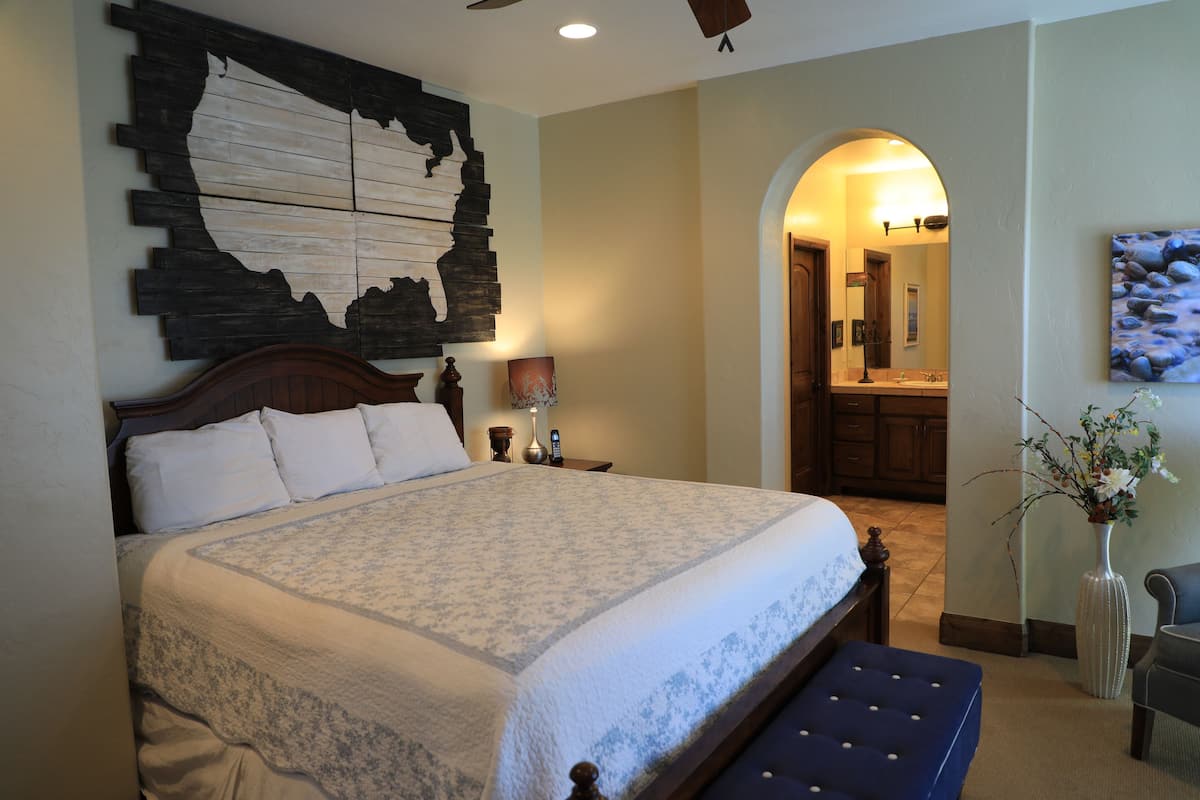 A bedroom with a large bed, a wooden headboard, a nightstand with a lamp, a vase with flowers, and a painting on the wall.