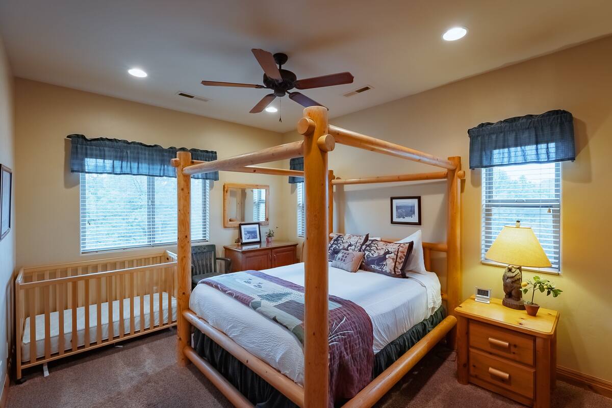 A four-poster bed with a wooden frame, a bedside table with a lamp, and a crib in a well-lit room with windows and a ceiling fan.