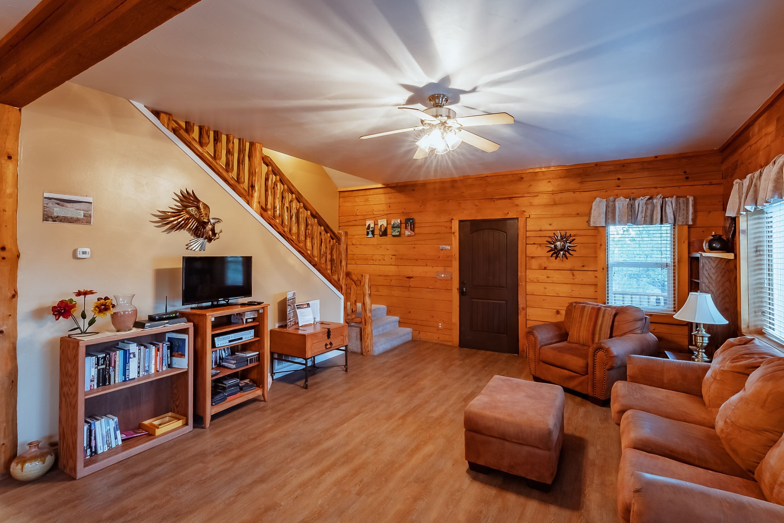 Ein gemütliches Blockhaus-Interieur mit einer Holztreppe, einem Fernseher, Bücherregalen, einer Couch und einem Deckenventilator.