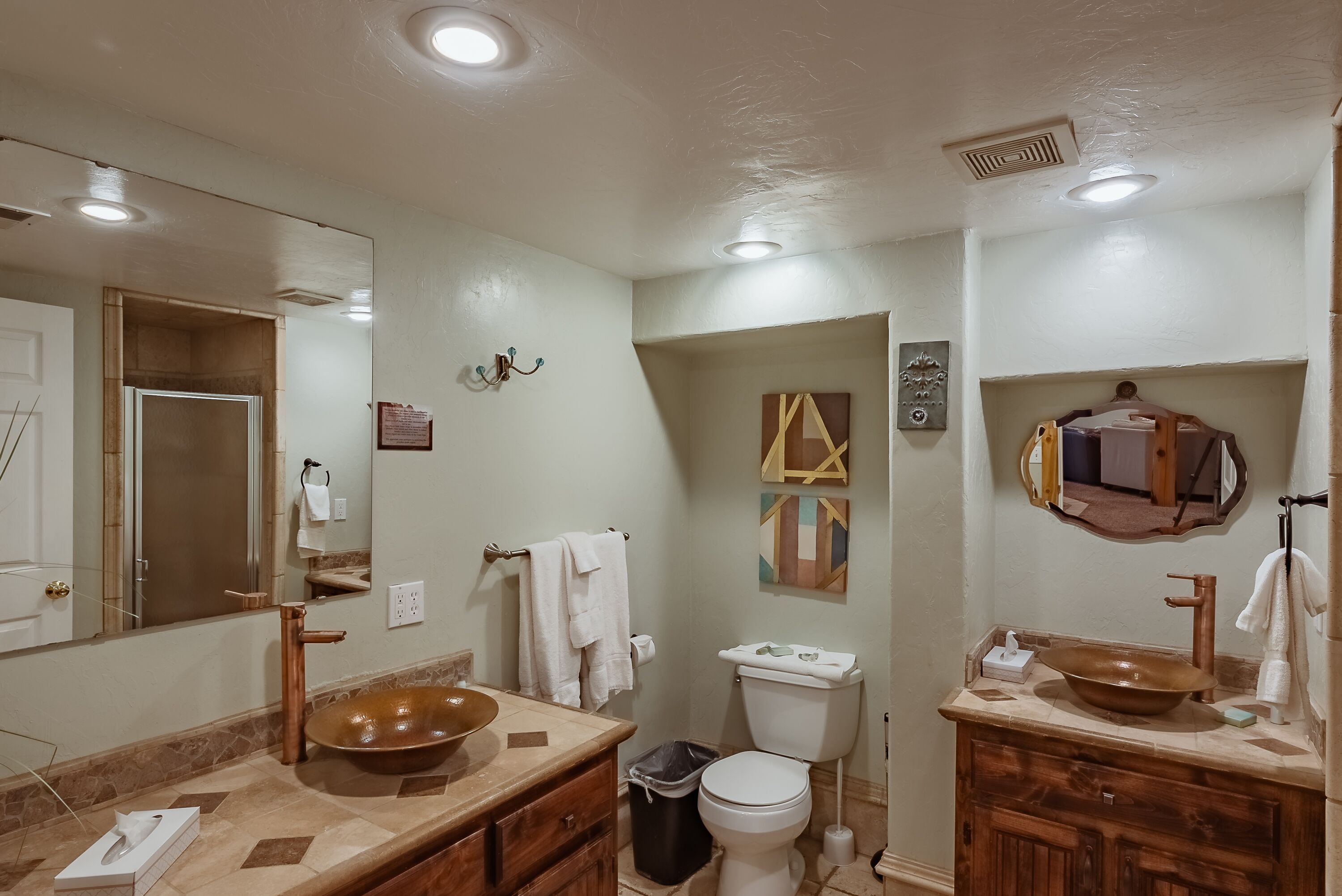 Un baño con un espejo grande, dos lavamanos con grifos de cobre, un inodoro y un cubo de basura.