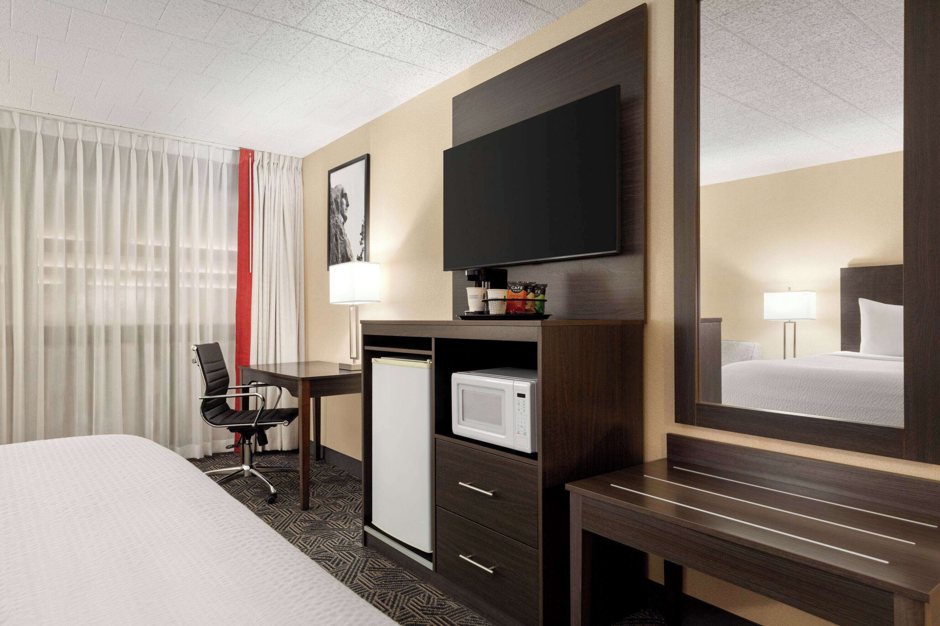 A hotel room with a bed, a desk with a microwave, a television, and a chair.