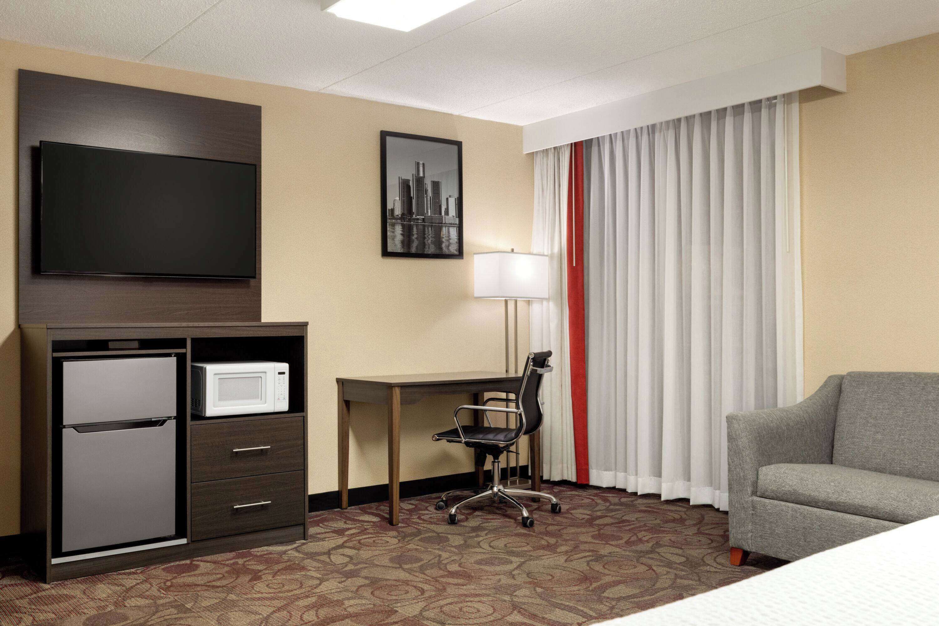 A hotel room with a bed, a desk with a microwave, a chair, a TV, and a cityscape picture on the wall.