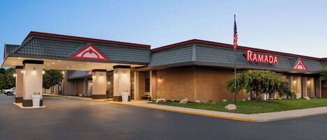 A Ramada hotel with a modern design, featuring a prominent red logo, a well-lit entrance, and a landscaped front yard.