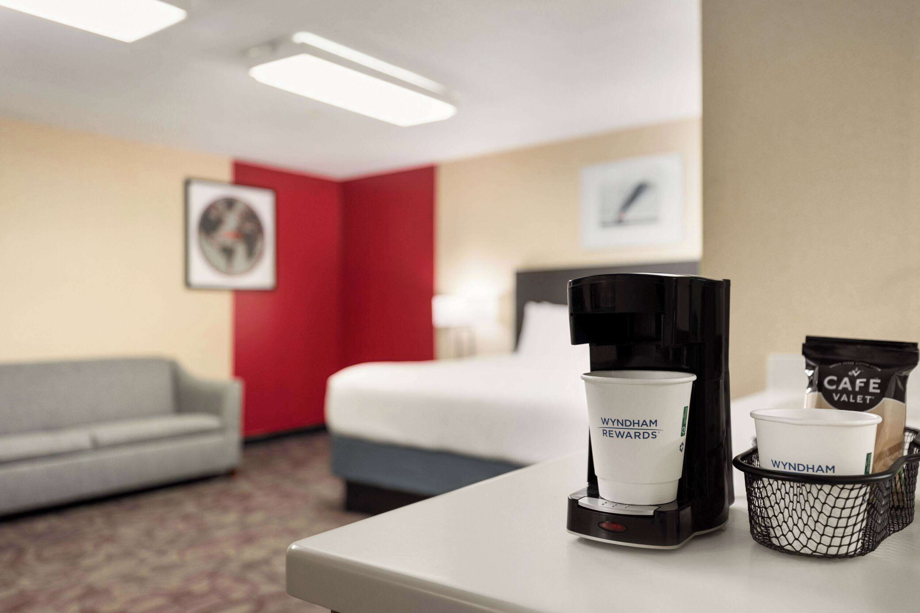 A hotel room with a bed, a coffee machine, and a basket with coffee cups and a bag labeled 'Café Valet'.