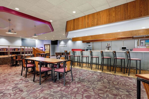 A restaurant with a bar area, multiple tables and chairs, and a patterned carpet. - Ramada by Wyndham Alpena (Alpena)