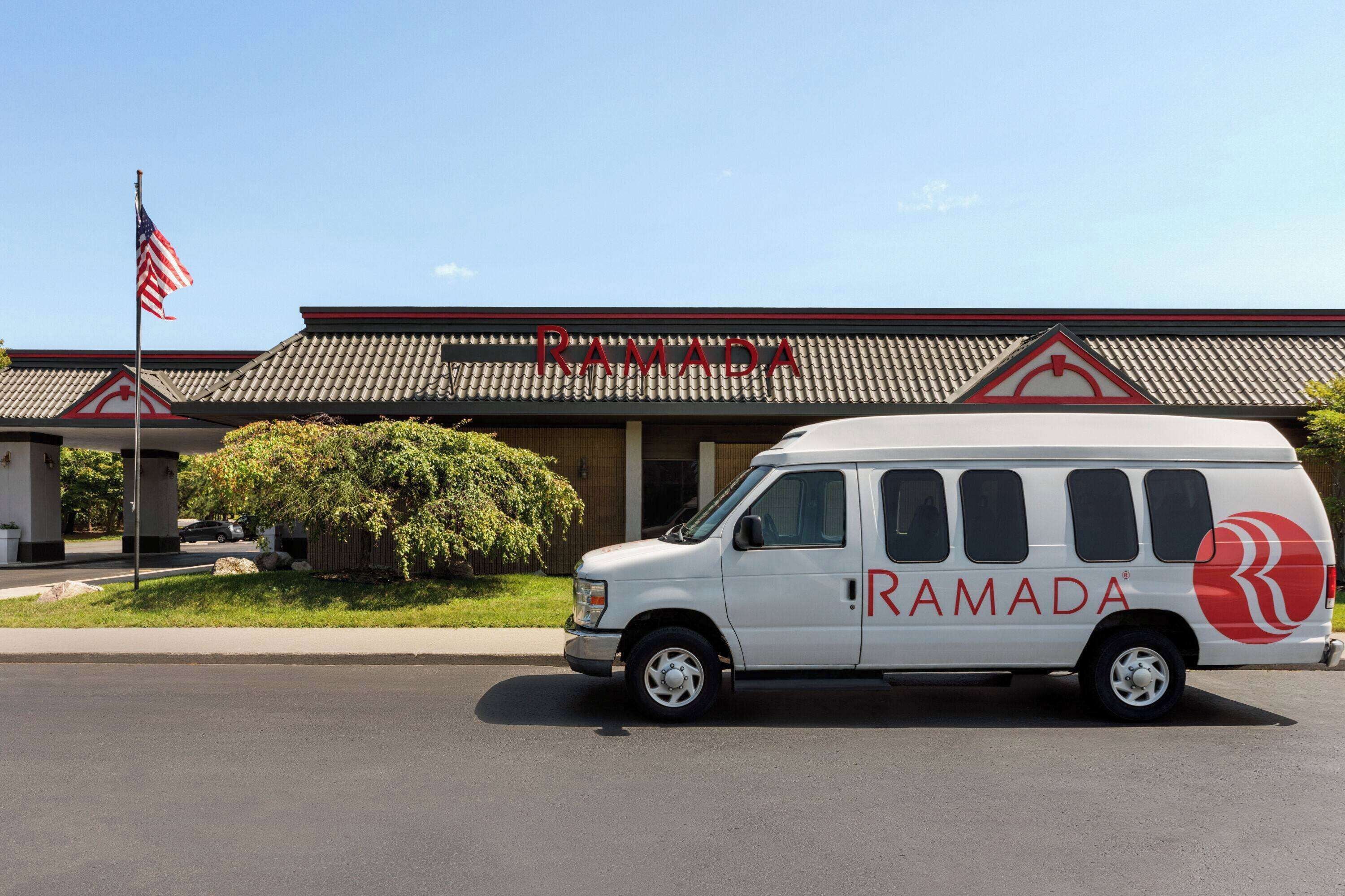 Ein weißer Van mit dem Schriftzug "RAMADA" und einem roten Logo steht vor einem Gebäude mit einem Ziegeldach und einer Fahnenstange mit der US-amerikanischen Flagge.