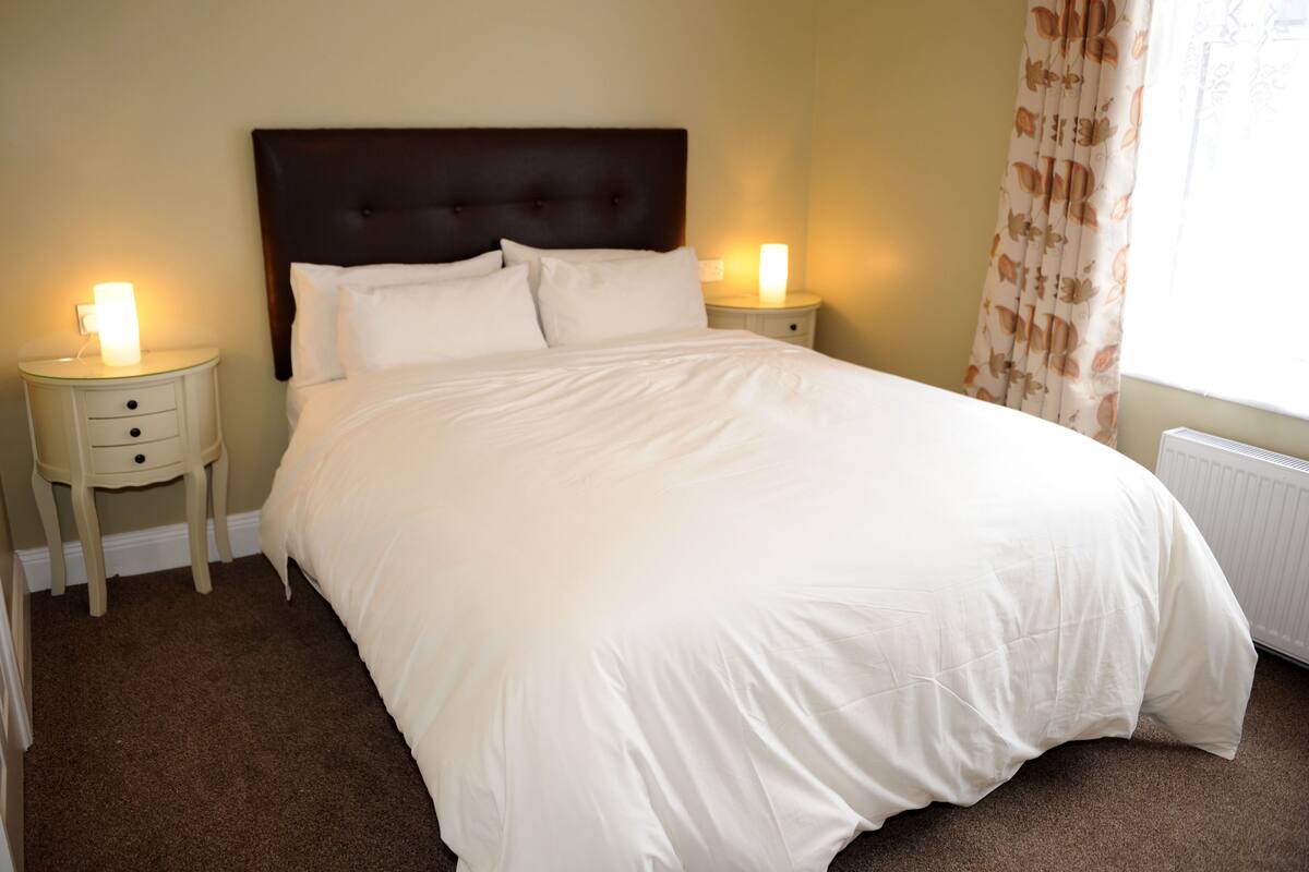Cama bem arrumada com lençóis brancos em quarto com paredes bege e cabeceira escura. Há duas mesas de cabeceira com abajures e uma janela com cortinas.