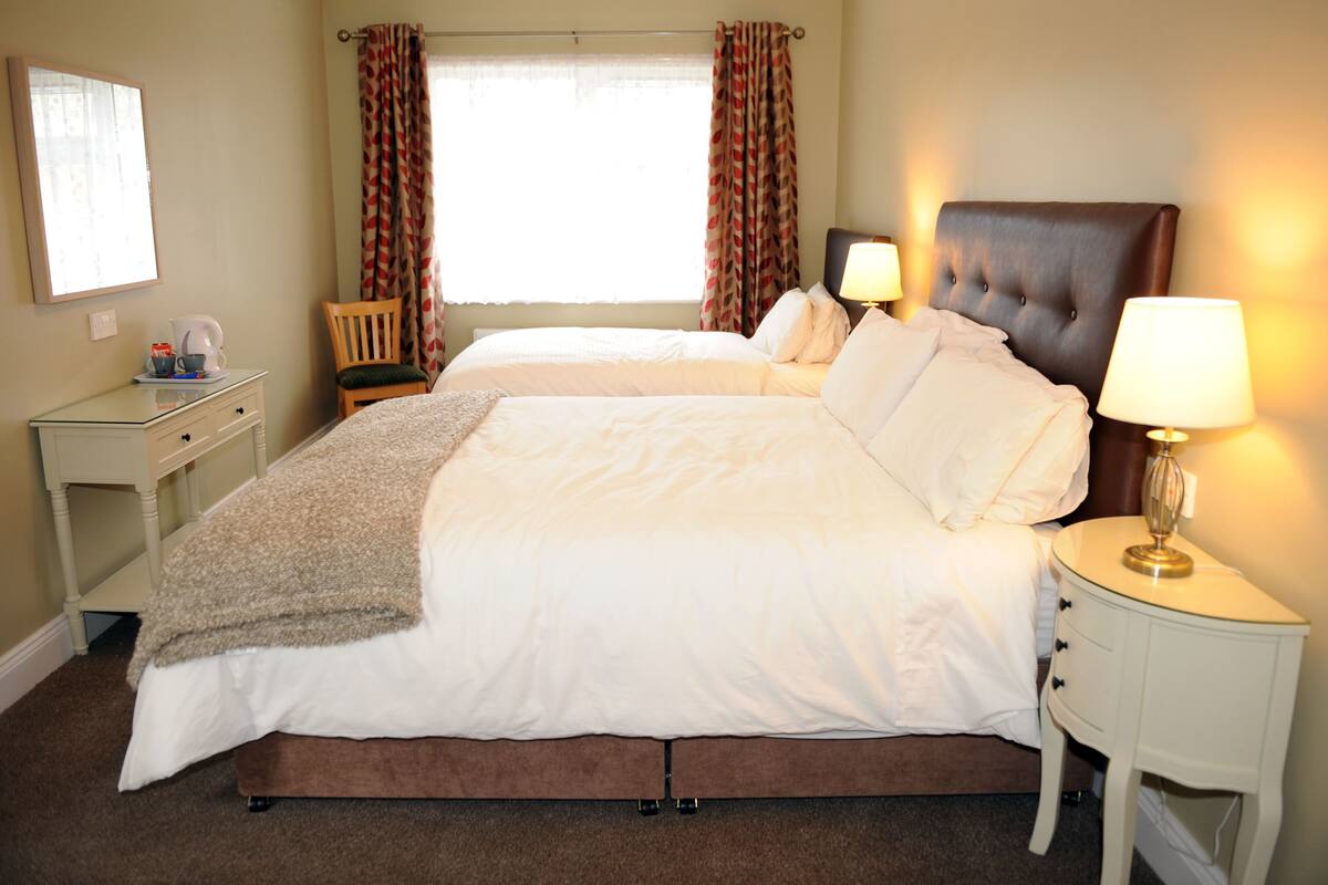 Quarto de hotel com duas camas, mesa de cabeceira com abajur, cadeira e janela com cortinas.