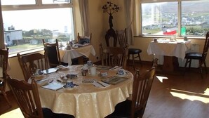 Un comedor con varias mesas y sillas, ventanas amplias que ofrecen vistas a un pueblo y montañas, y un jarrón decorativo sobre una de las mesas.