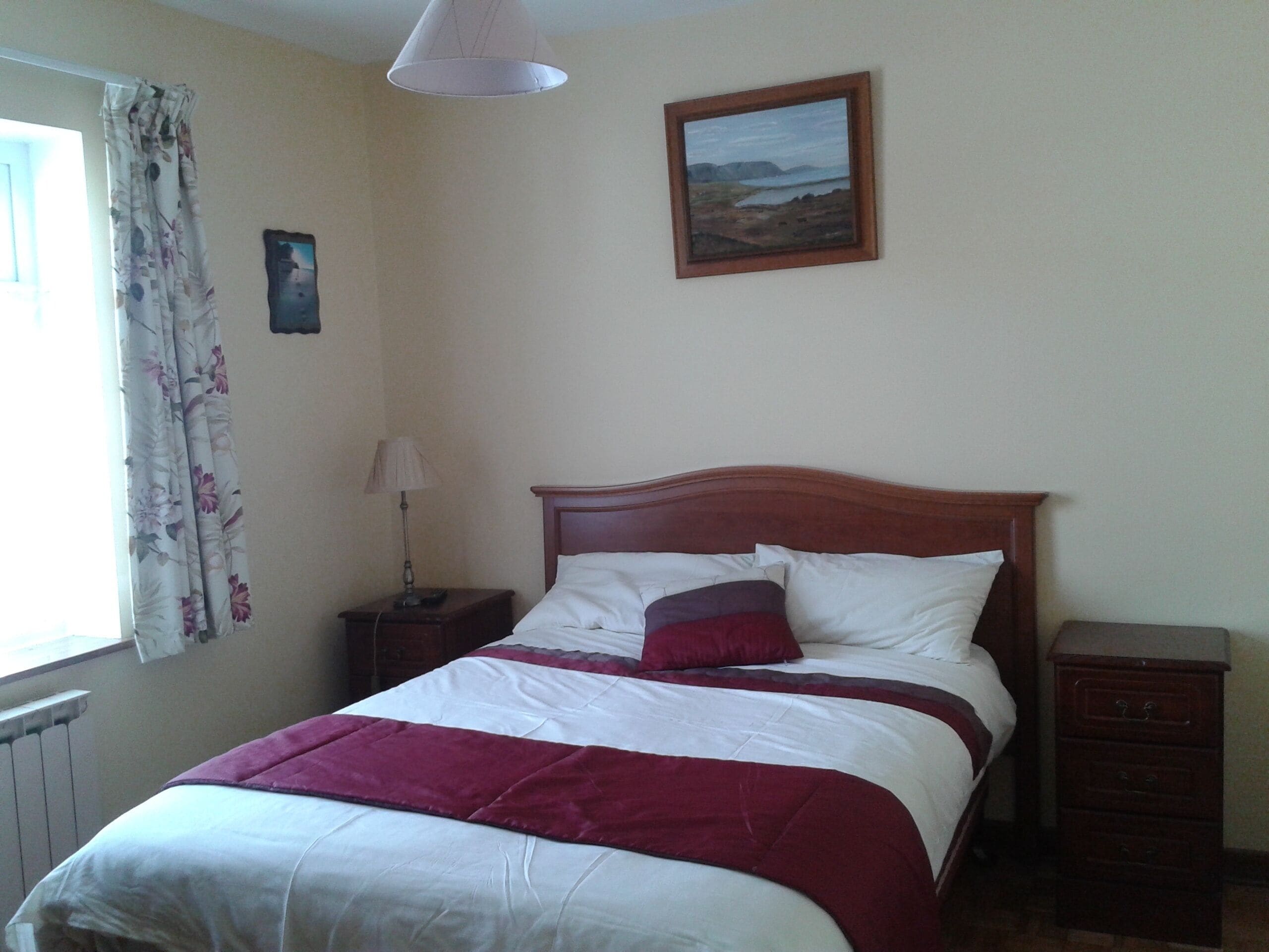 A bedroom with a bed, a nightstand, a lamp, a dresser, a window with curtains, and a painting on the wall.