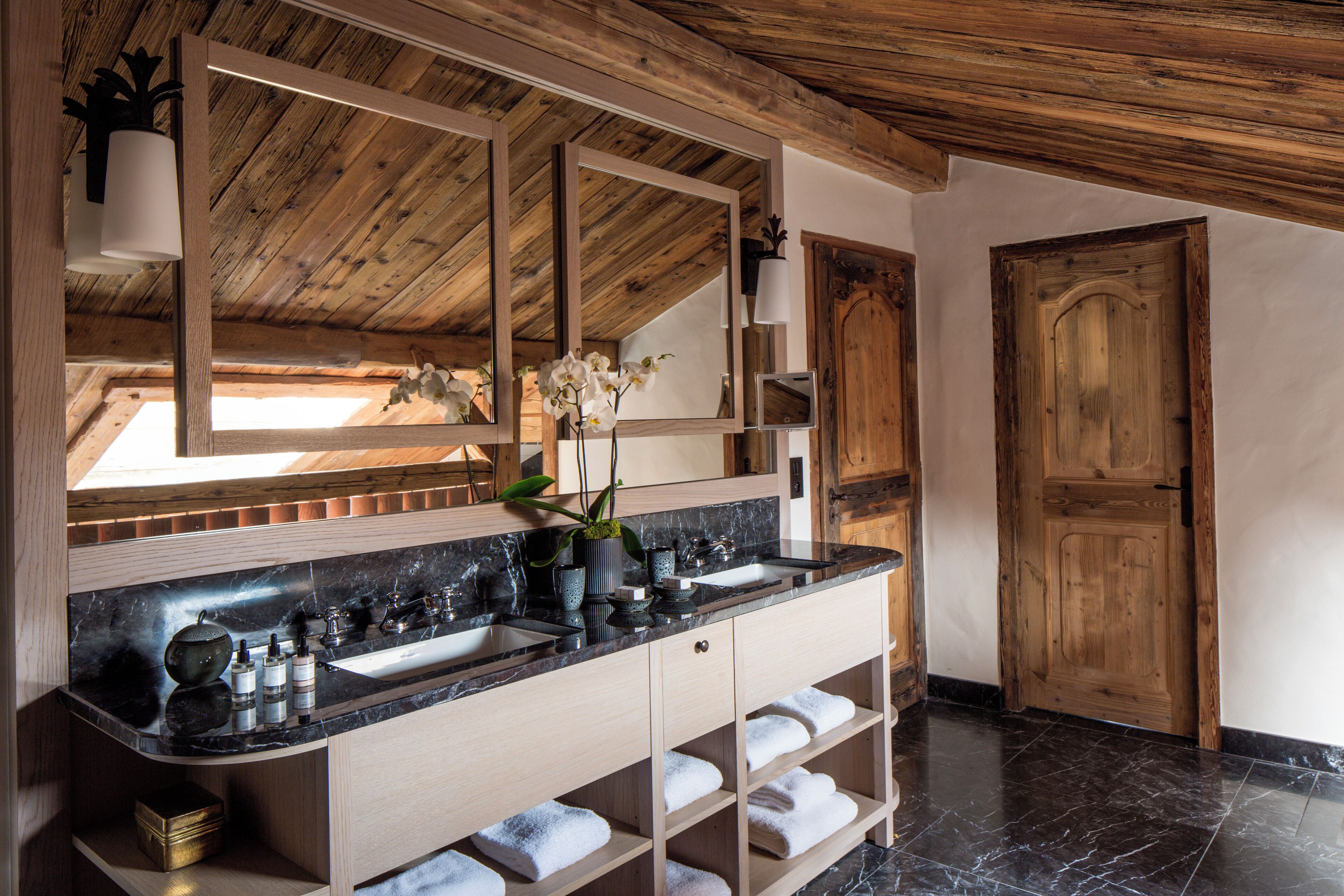 A modern bathroom with a wooden ceiling, a large mirror, a black marble countertop, and a wooden door.