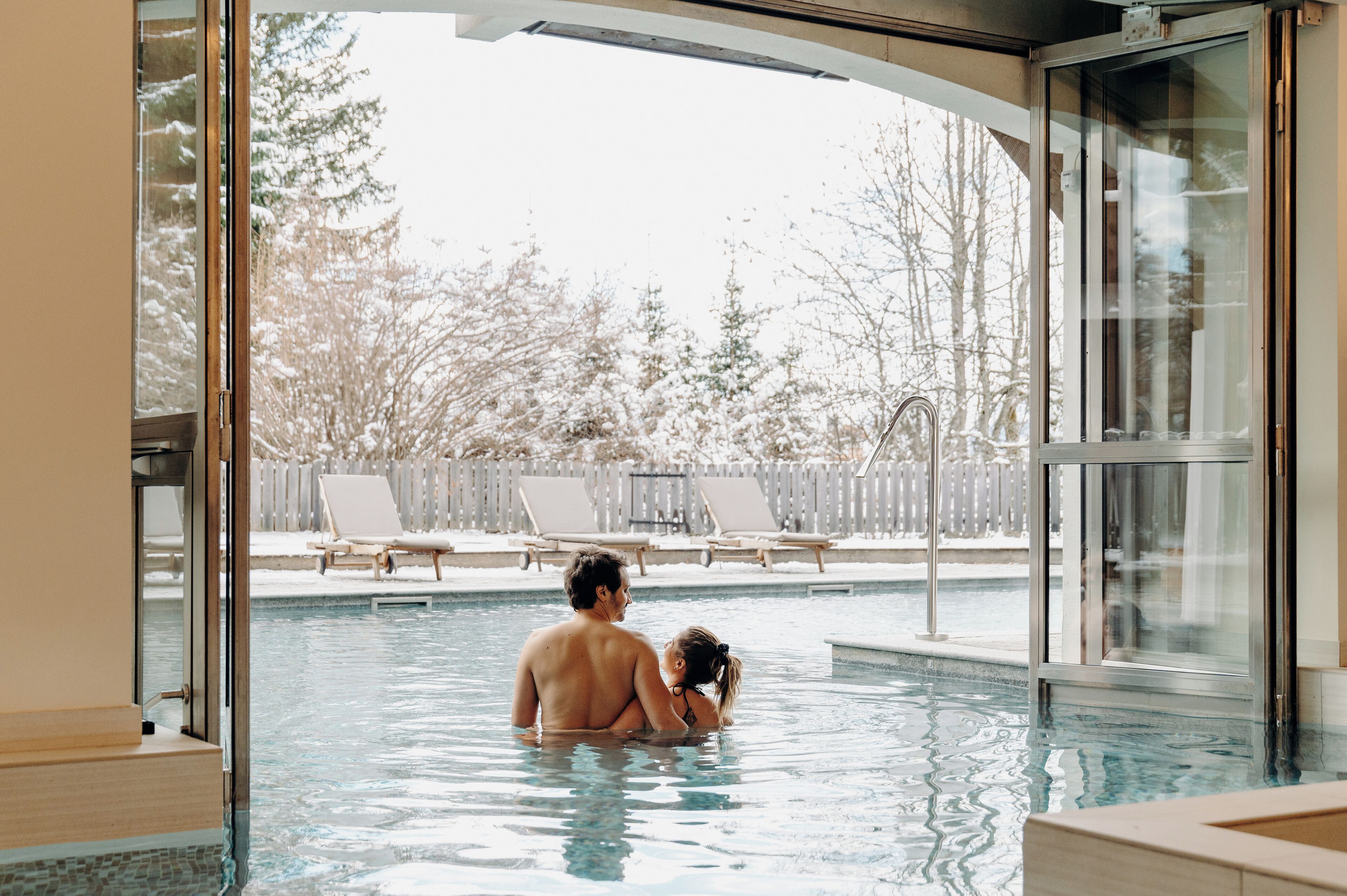A swimming pool with a view of a snowy landscape and a wooden deck with lounge chairs.