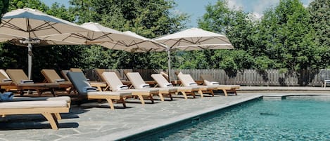 Zona de piscina con sillas de descanso y sombrillas, rodeada por una valla de madera y árboles.