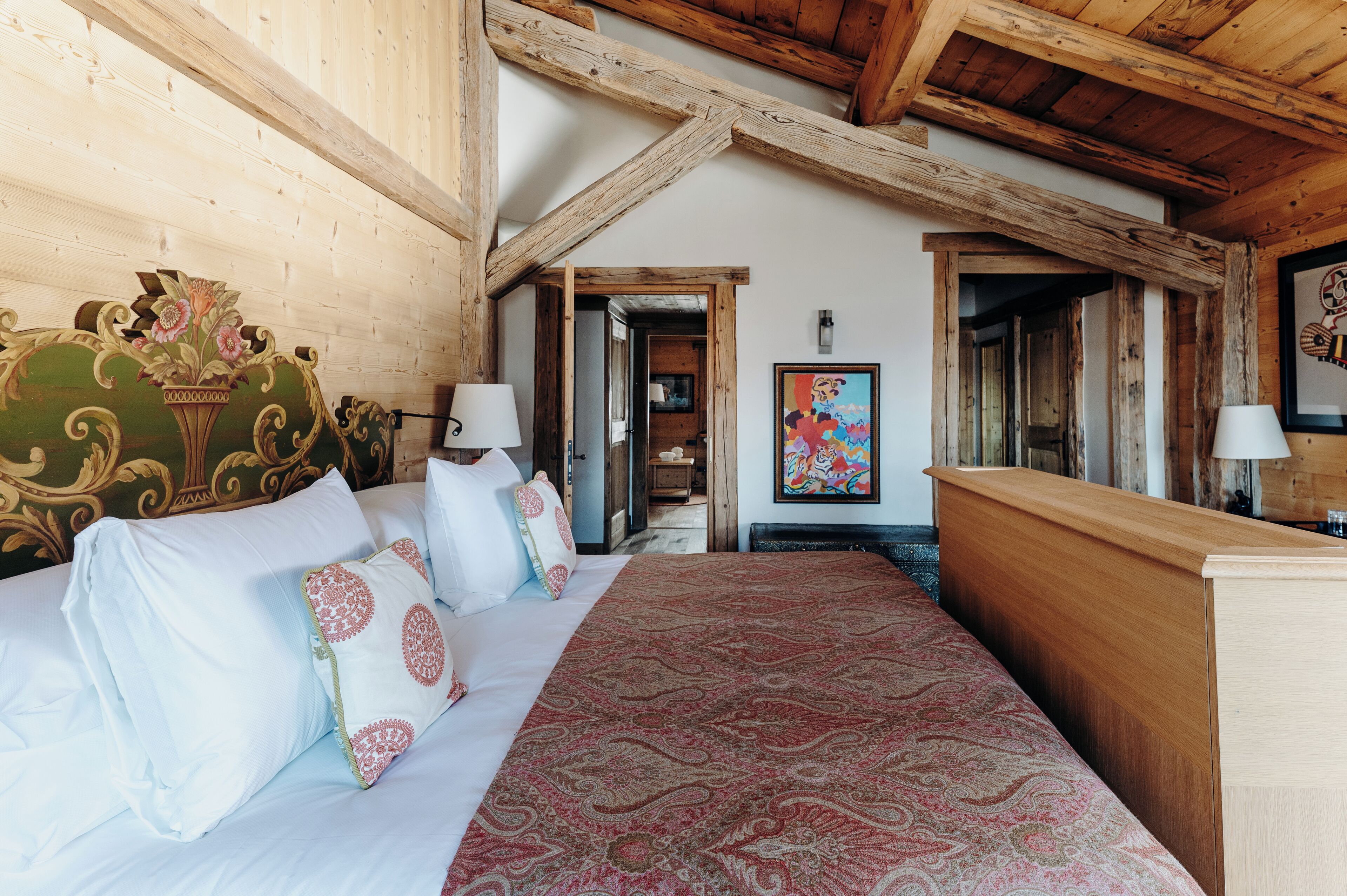 Un dormitorio con techo de madera, una cama con ropa de cama blanca y estampada, un aparador de madera y un cuadro colorido en la pared.