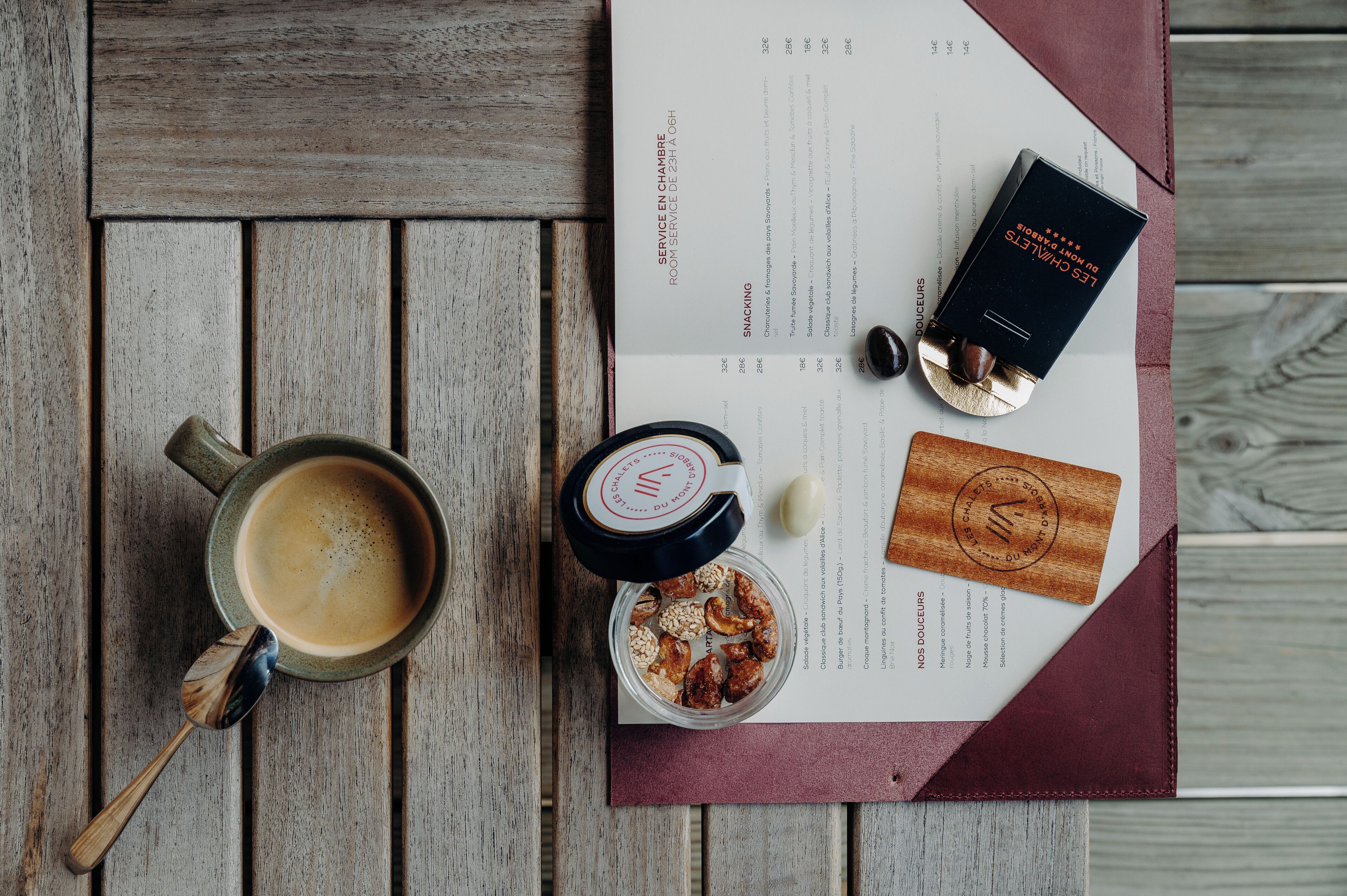 Eine Speisekarte mit Gerichten und Preisen, eine Kaffeetasse, ein Holzlöffel und eine kleine Nussdose auf einem Holztisch.