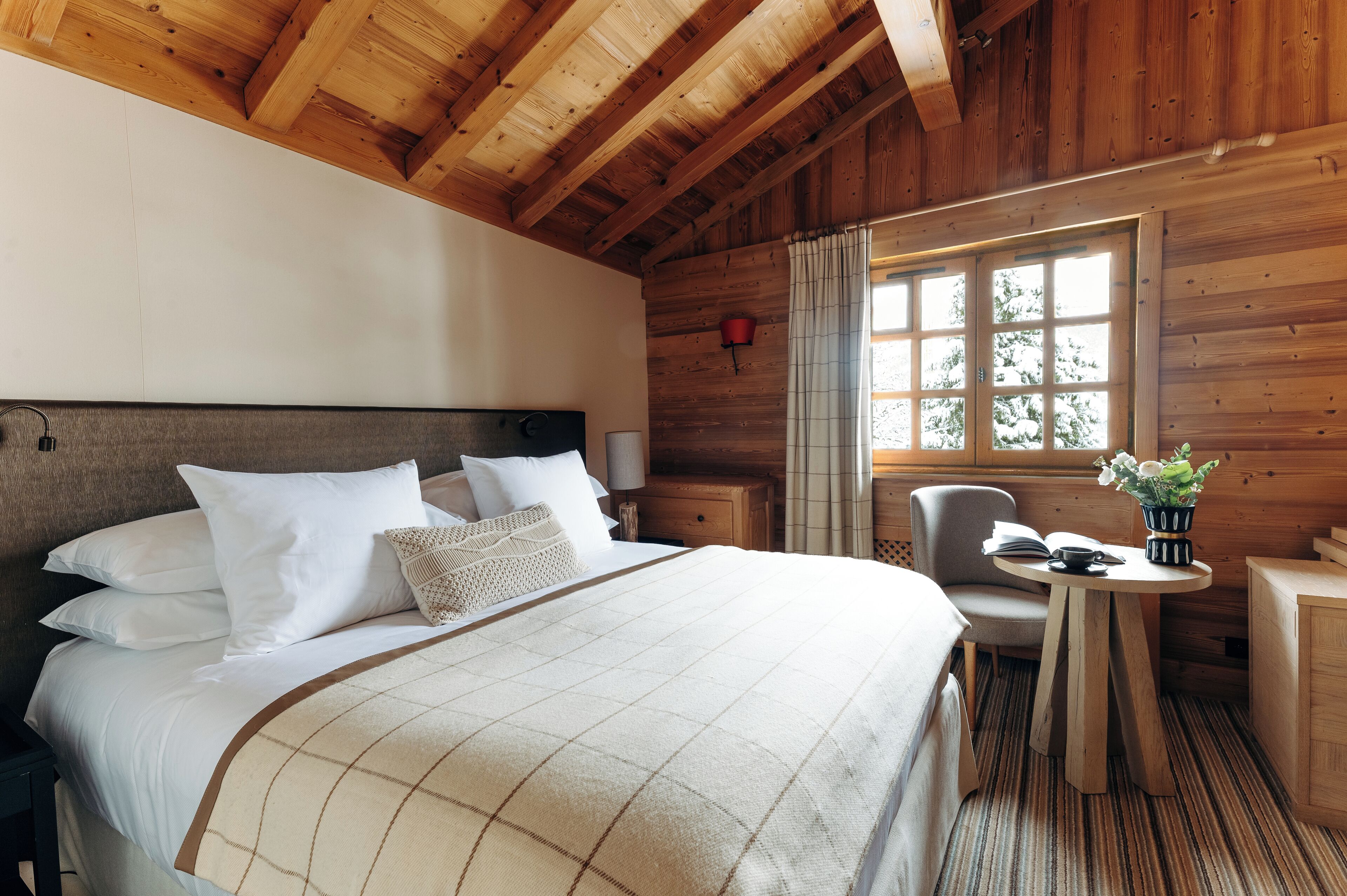 A wooden-paneled room with a large bed, a small table with a plant, and a window with curtains.