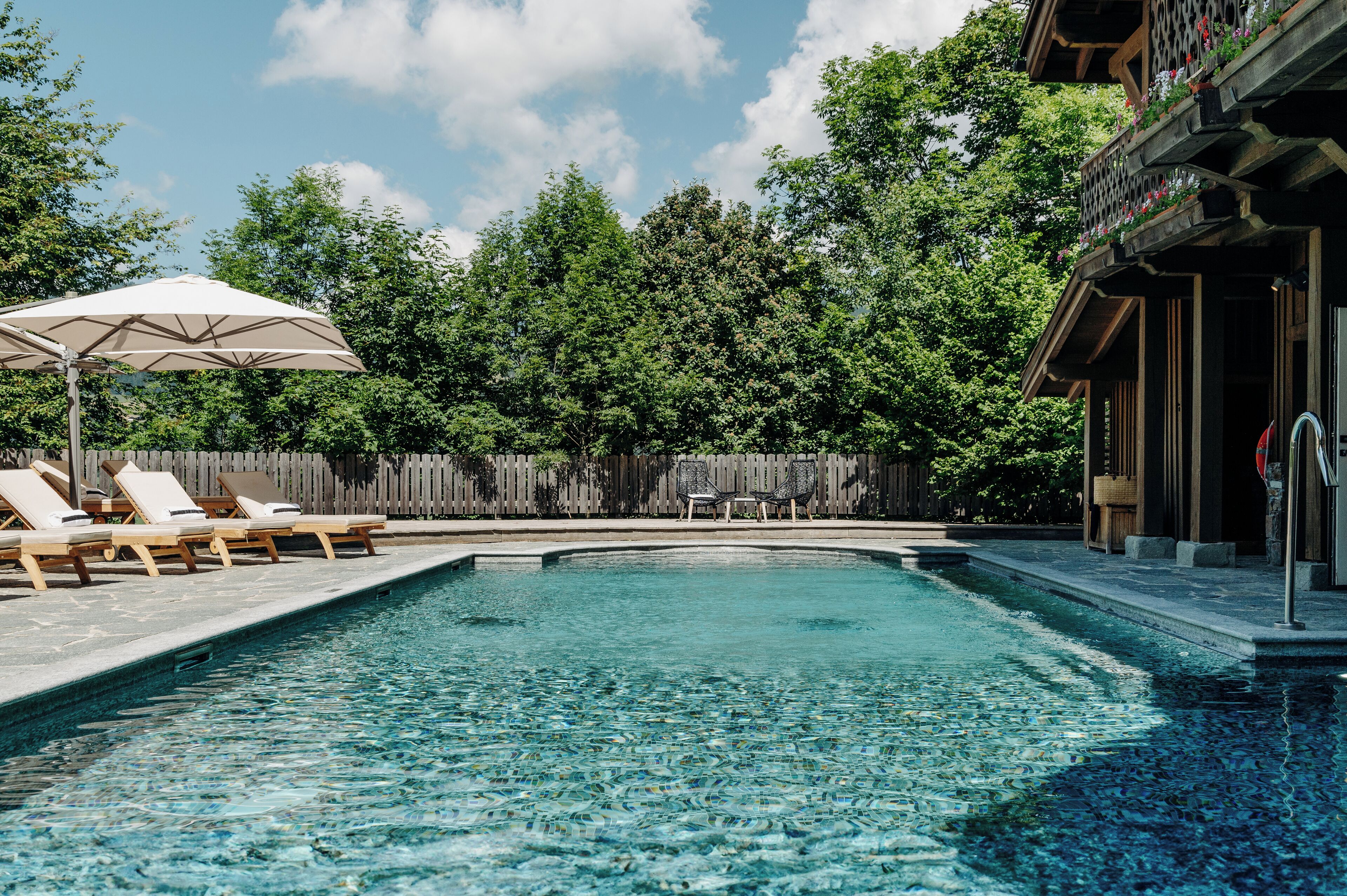 A swimming pool with lounge chairs and an umbrella, surrounded by a wooden fence and trees.