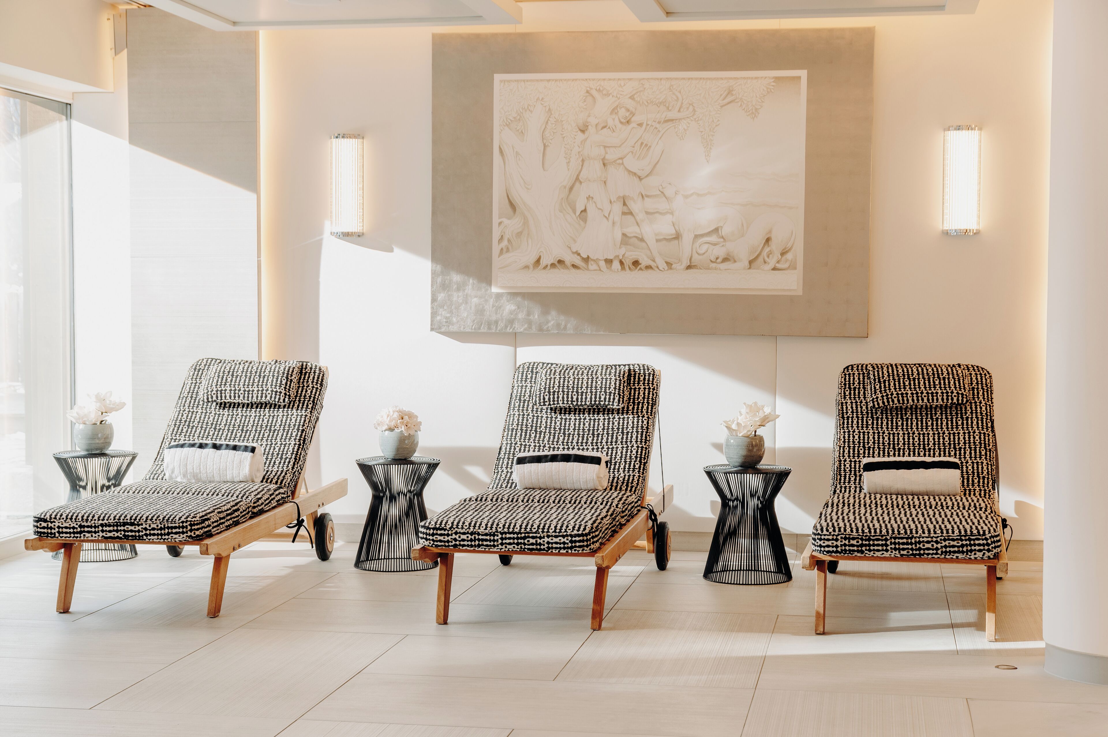 Three patterned lounge chairs with wooden frames and black metal side tables, arranged in a sunlit room with a large wall-mounted sculpture.