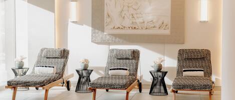 Three patterned lounge chairs with wooden frames and black metal side tables, arranged in a sunlit room with a large wall-mounted sculpture.