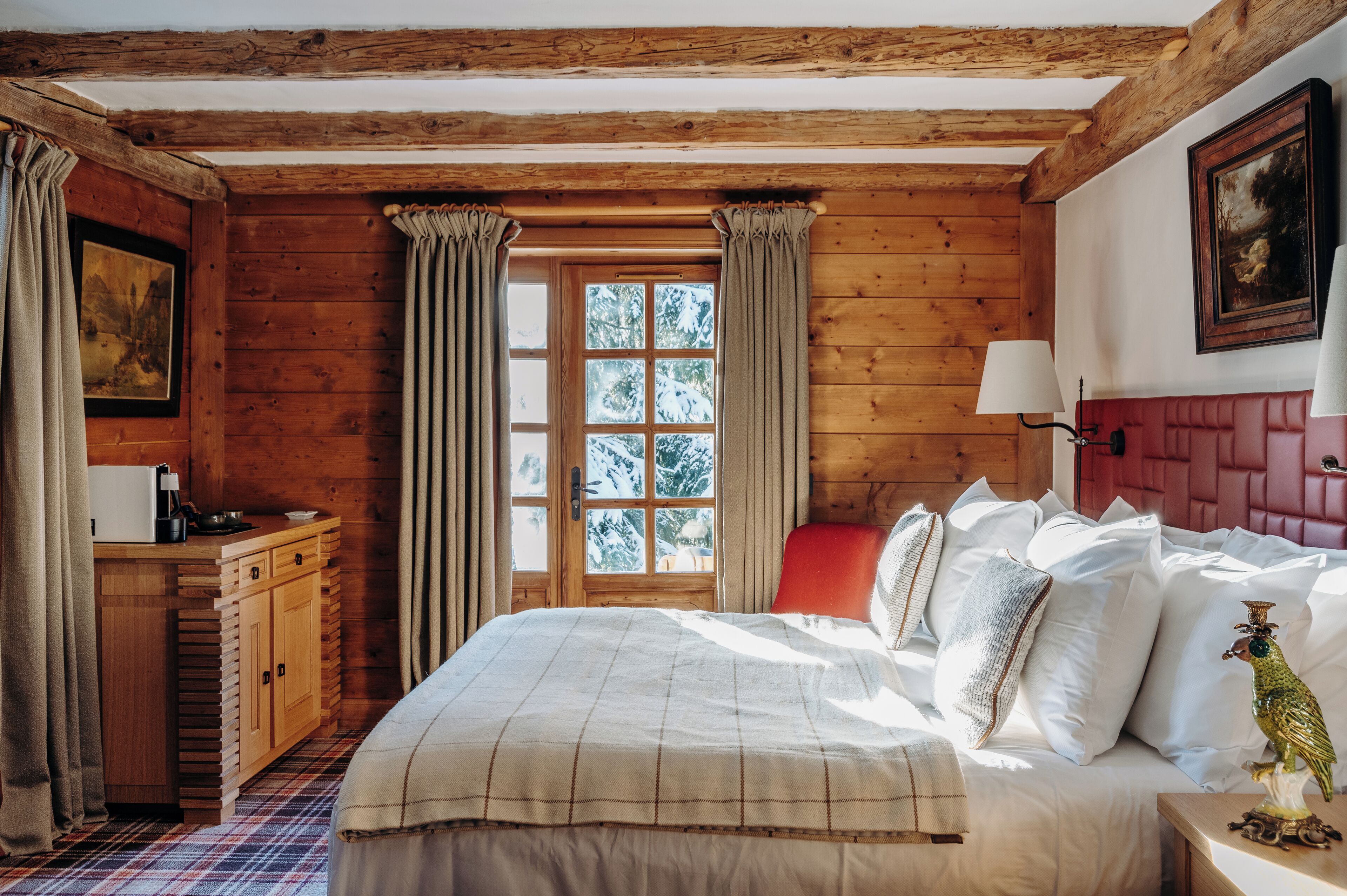 A wooden cabin-style bedroom with a bed, a dresser, a window with curtains, and a painting on the wall.