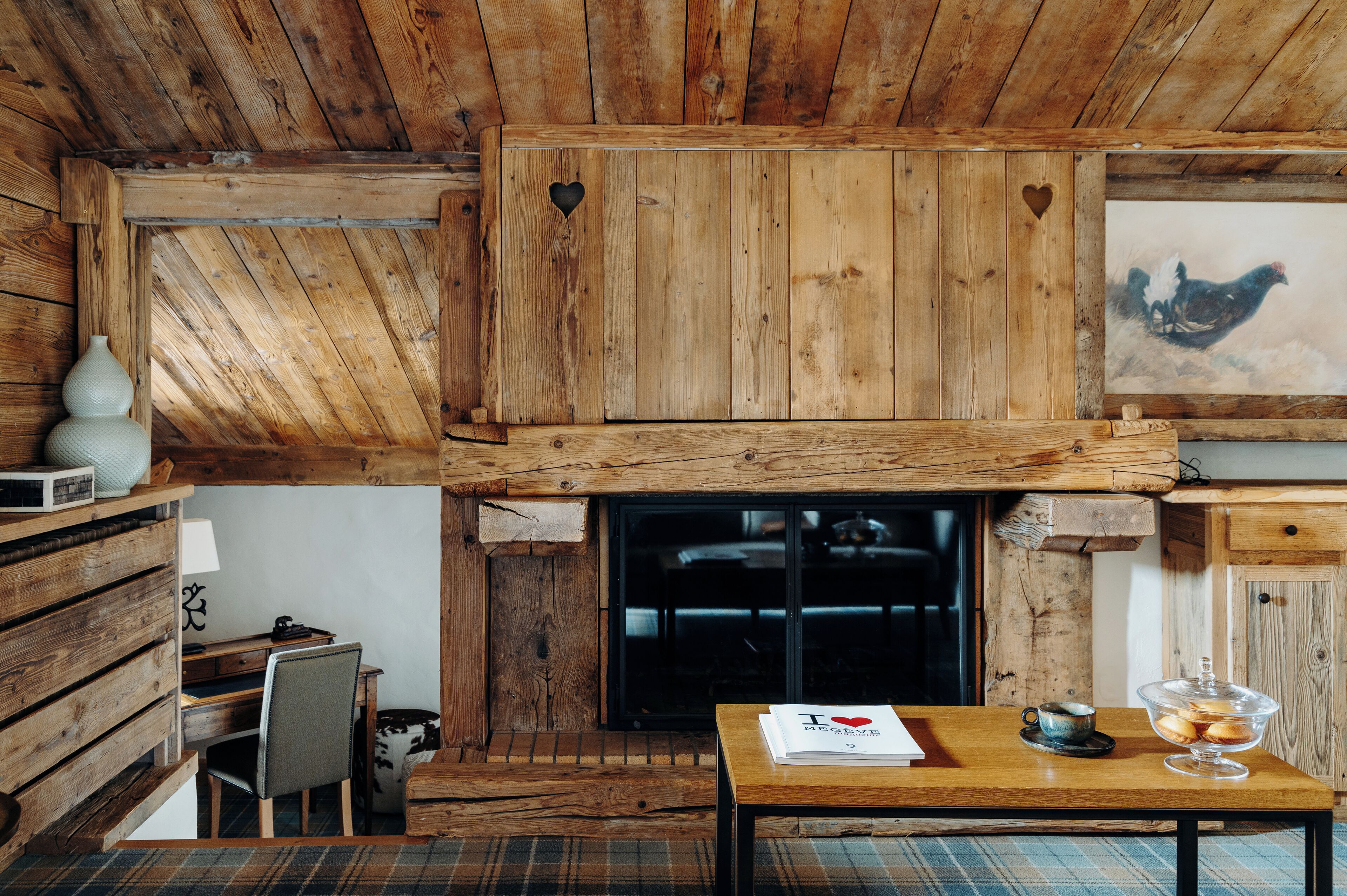 A rustic wooden interior with a fireplace, a wooden desk, and a dining table with a clear glass cover.