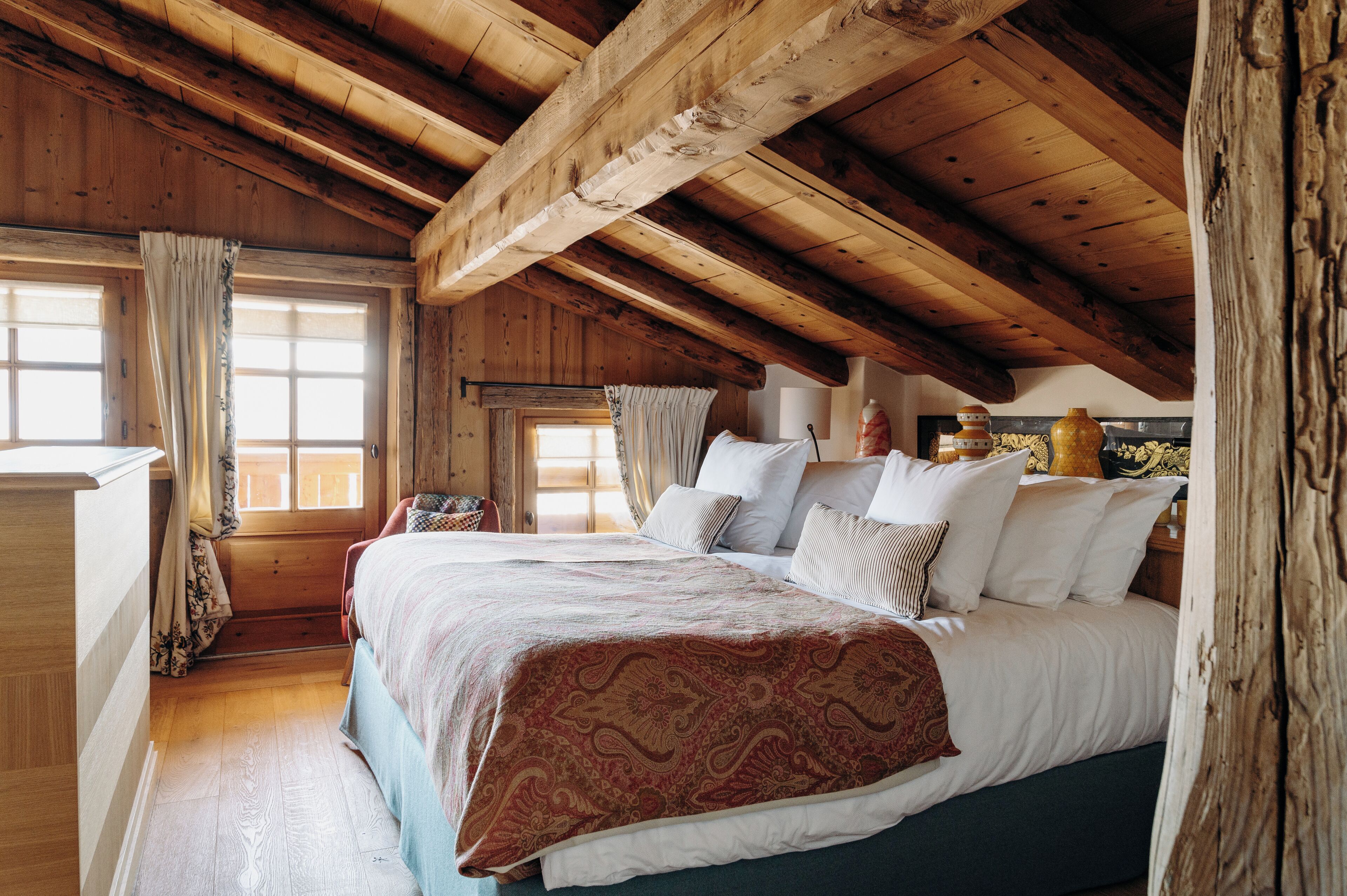 A bedroom with a wooden ceiling, a large bed with multiple pillows, a dresser, and a window with curtains.