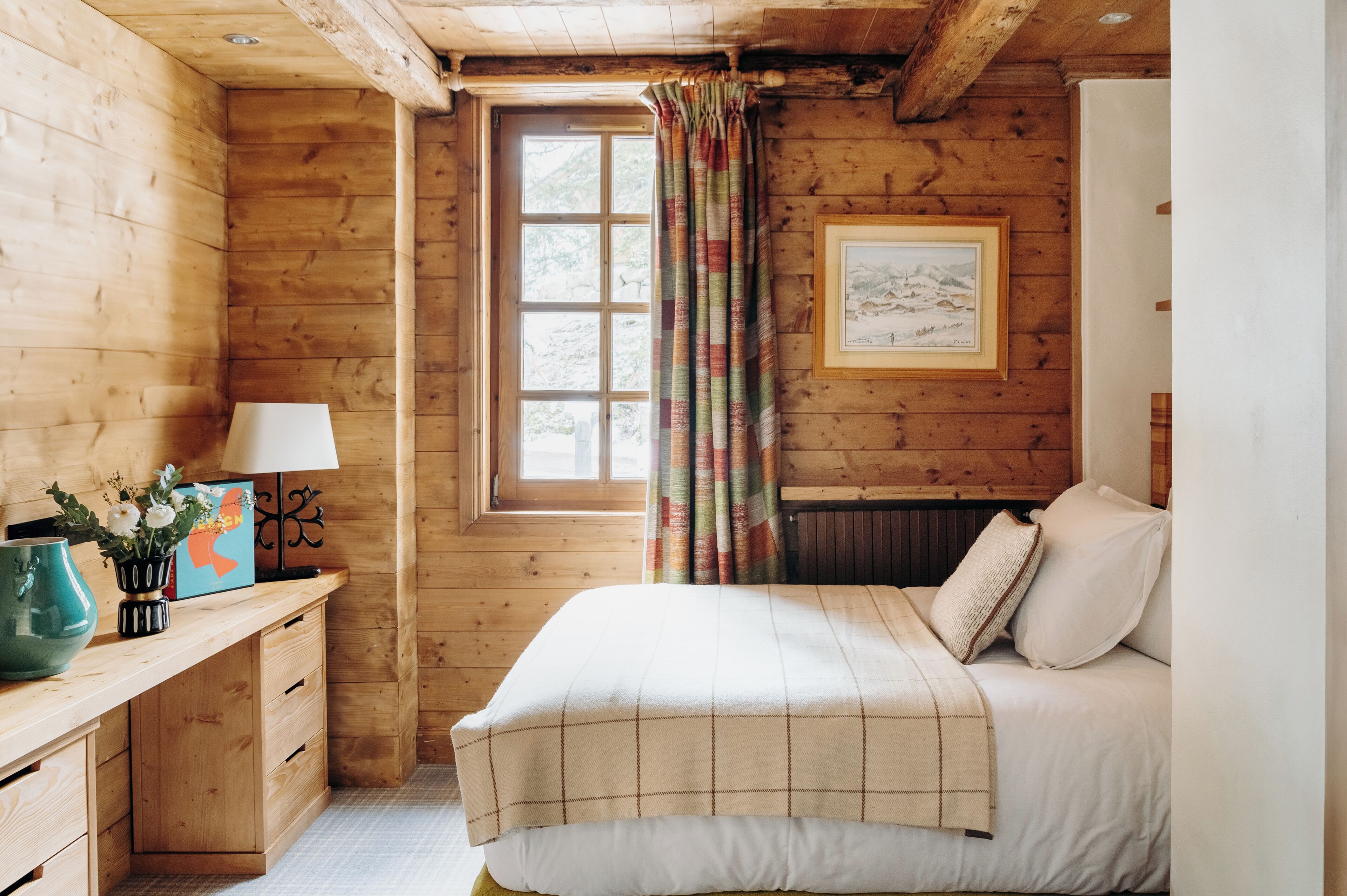 A cozy wooden room with a bed, a desk with a lamp, and a window with curtains.