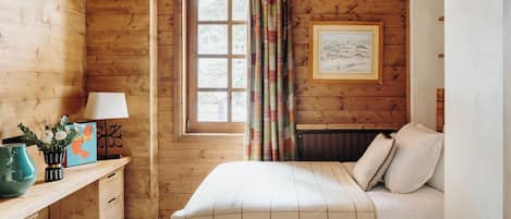 A cozy wooden room with a bed, a desk with a lamp, and a window with curtains.