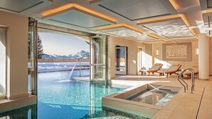 Area piscina interna moderna con pareti di vetro trasparente, poltrone relax e vista sulle montagne innevate.