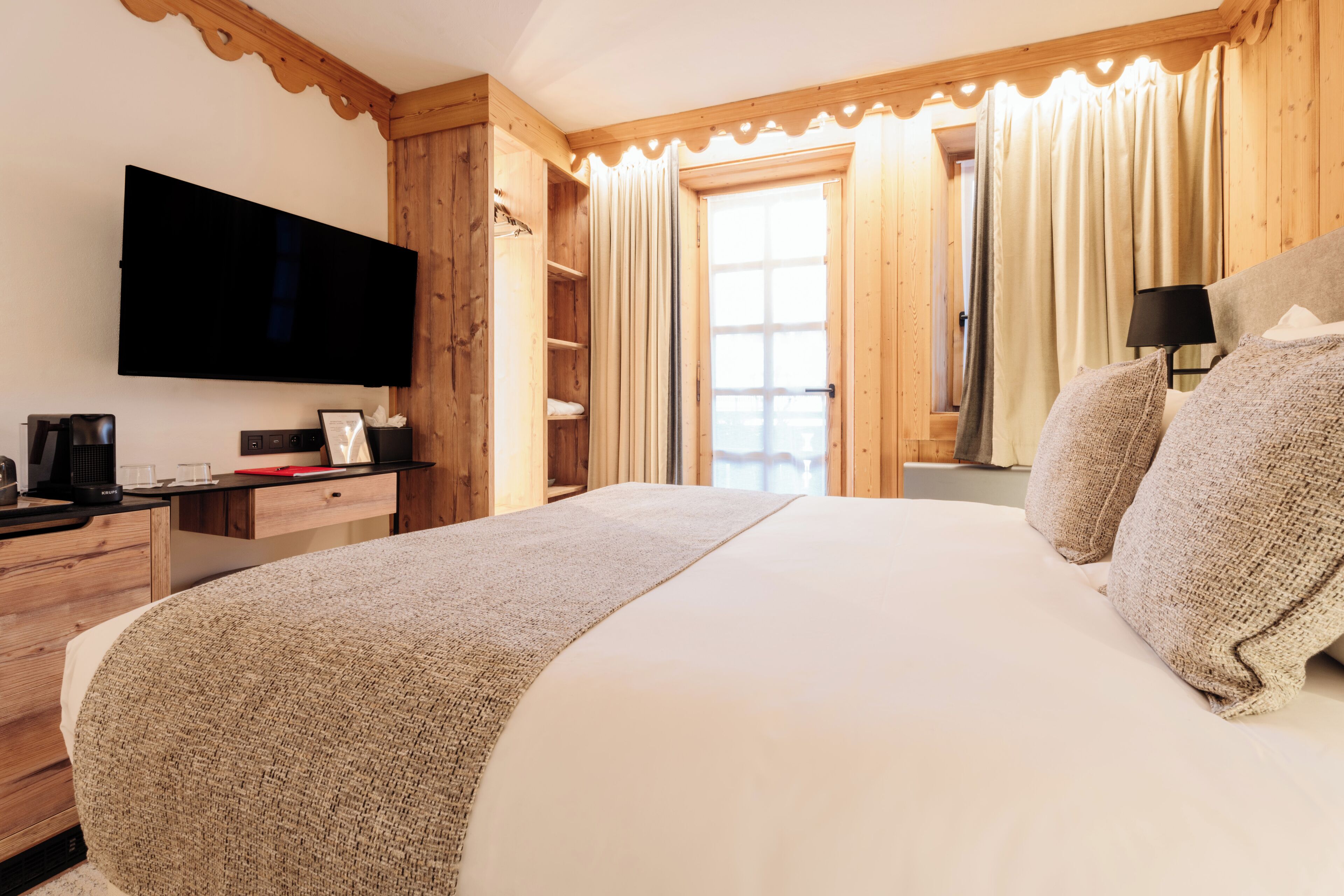 Una habitación de hotel con una cama grande, muebles de madera, un televisor de pantalla plana, un escritorio con cafetera y una ventana con cortinas.