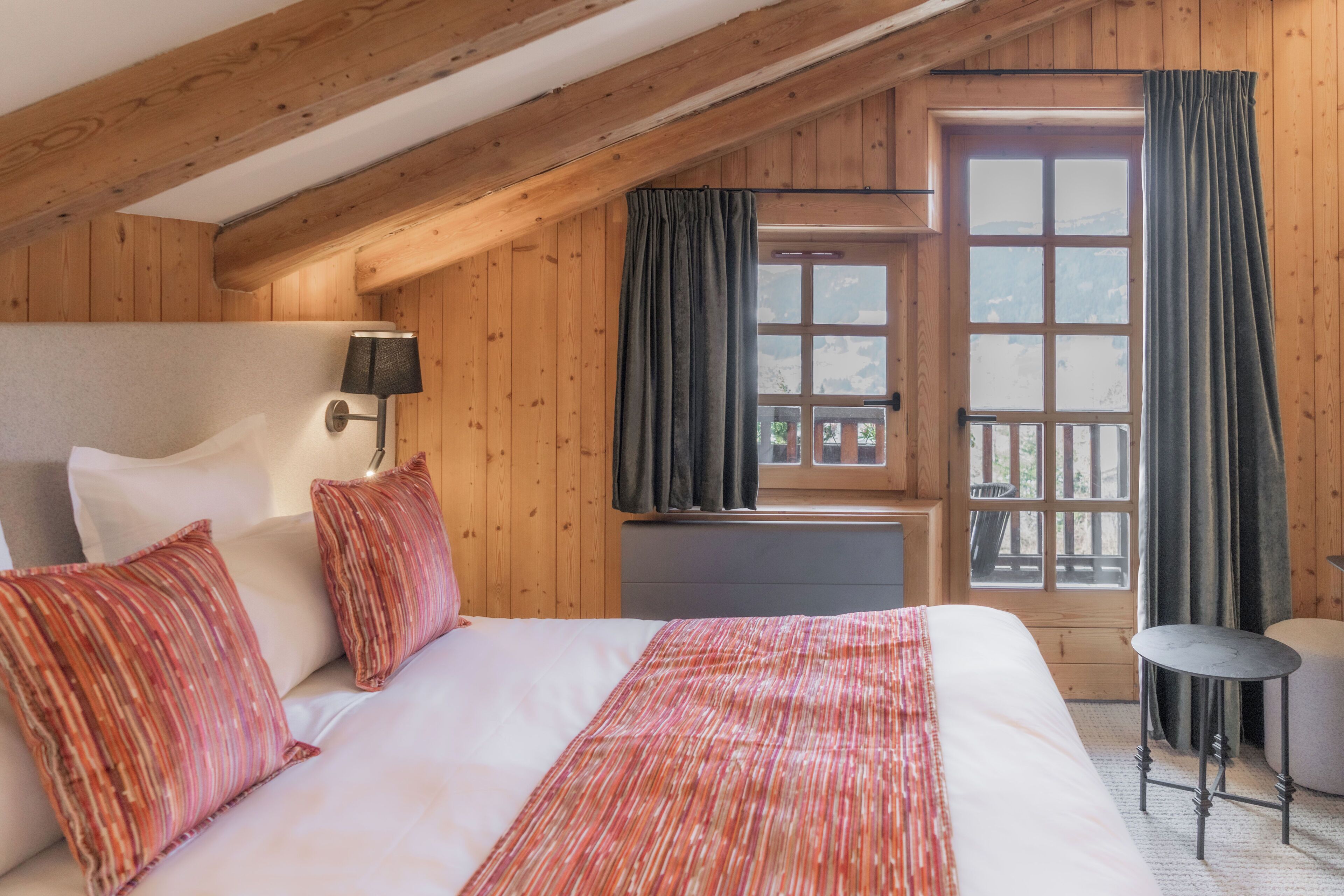 Una habitación acogedora con techo de madera, una cama grande con almohadas a rayas, una mesita de noche y vistas a un balcón por la ventana.
