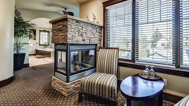 A cozy living room with a stone fireplace, a striped armchair, a round table with a vase, and large windows with blinds. - Westmount River Inn (Calgary)