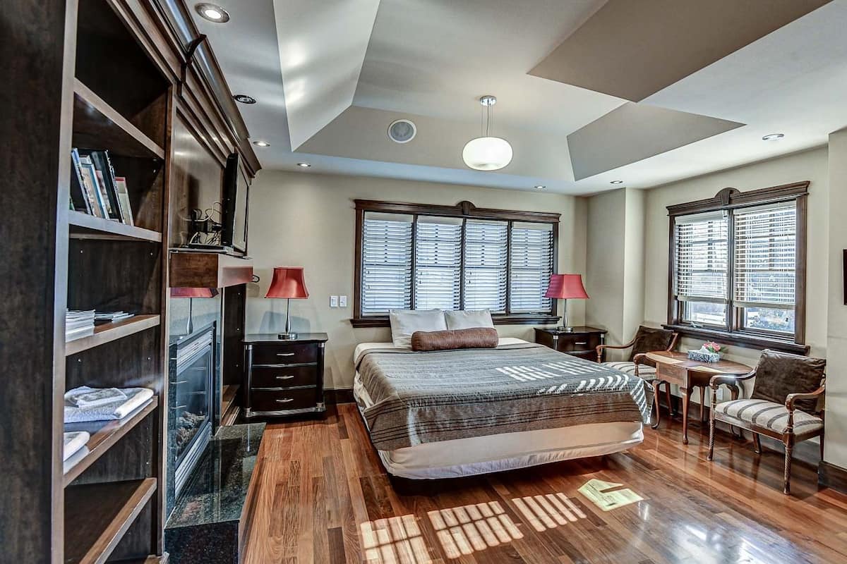 A bedroom with a large bed, wooden floor, built-in bookshelf, two bedside tables with lamps, a small table with chairs, and large windows with blinds.