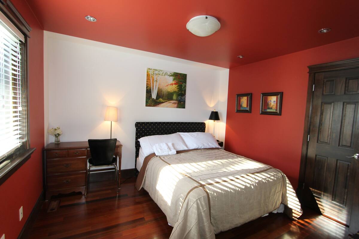 A bedroom with a bed, a desk, and a chair. There is a painting on the wall and a door to the right.
