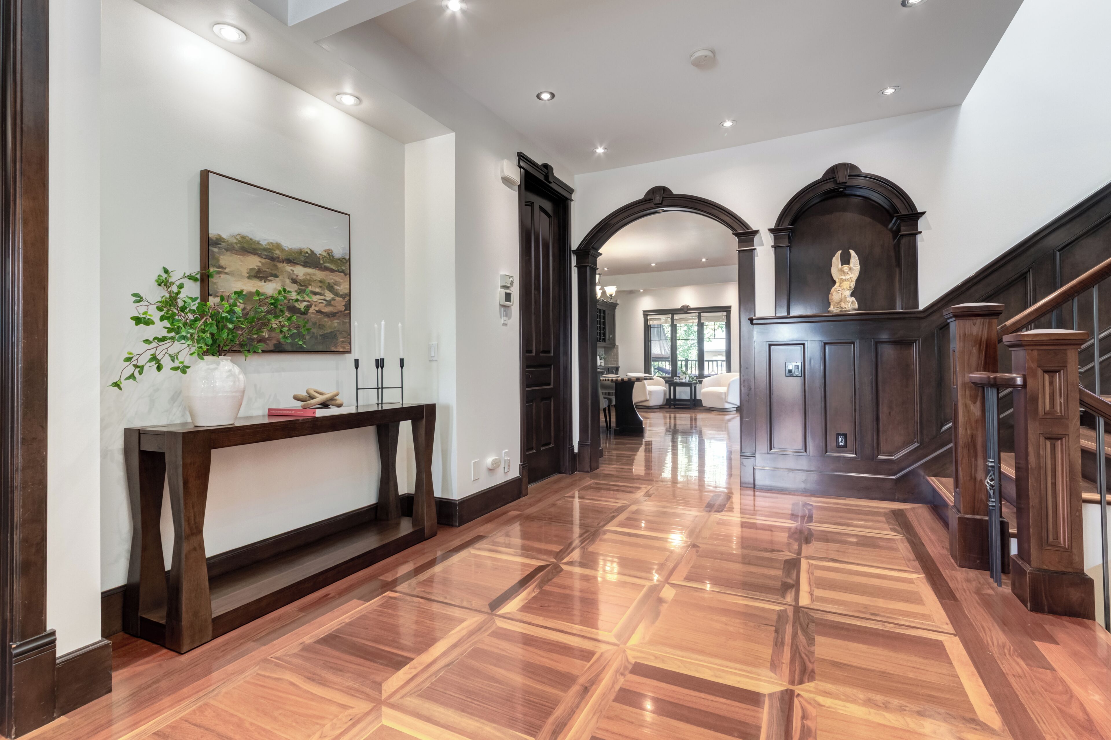 A polished wooden floor, a console table with a vase and a painting, and a statue in a dark wooden alcove.