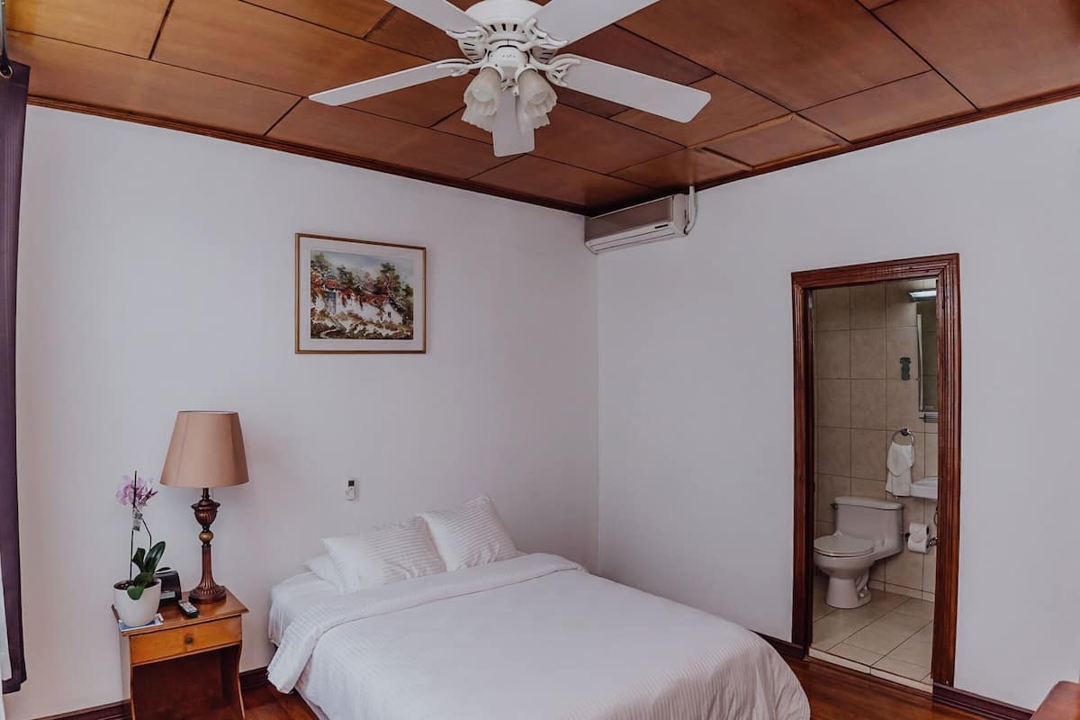 A bedroom with a bed, a nightstand with a lamp, a ceiling fan, a framed picture on the wall, and a bathroom visible through an open door.