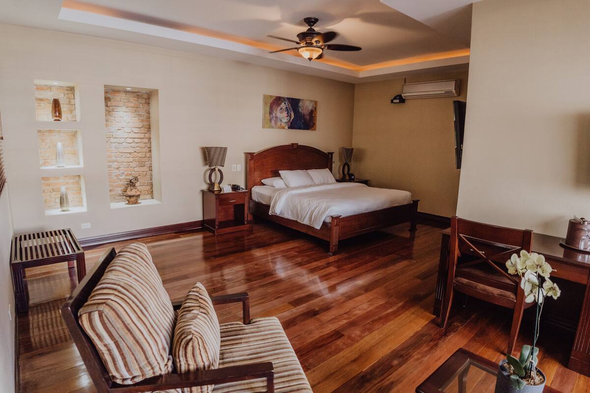 Un dormitorio con una cama de madera, una silla de madera, una mesita pequeña y un ventilador de techo.