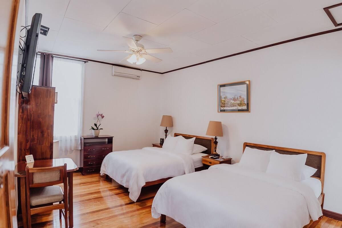 Habitación de hotel con dos camas, un escritorio, una silla, un ventilador de techo y una ventana con cortinas.