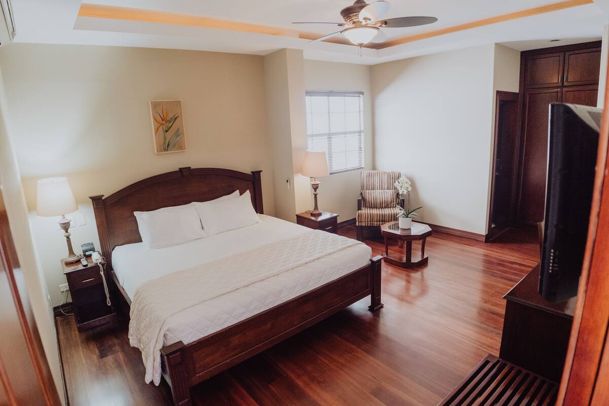 Una habitación de hotel con una cama grande, televisor, una silla, una lámpara y un ventanal.