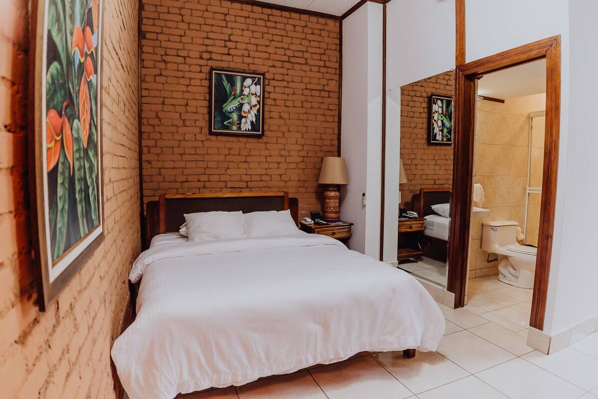 Un dormitorio con pared de ladrillo, cama con ropa de cama blanca, mesita de noche con lámpara y baño visible a través de una puerta abierta.