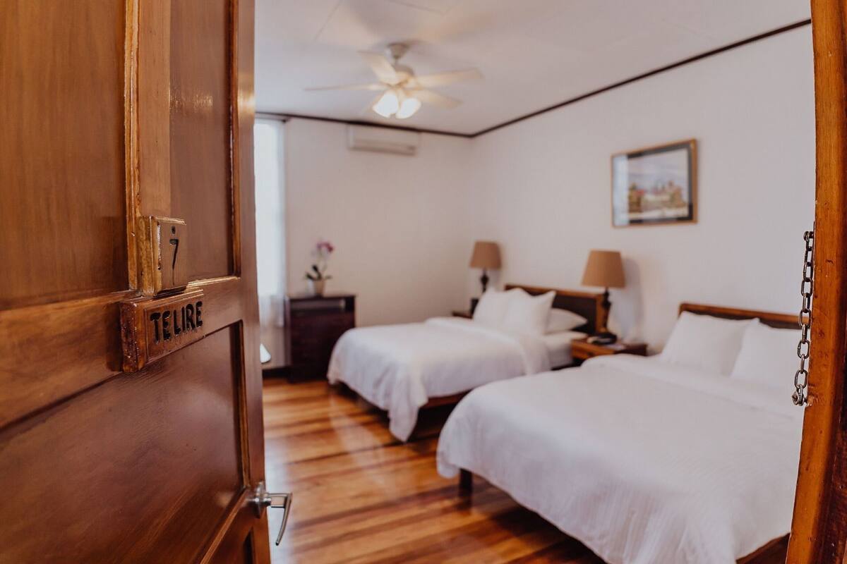 Habitación de hotel con dos camas, una puerta de madera con cerradura numérica, un ventilador de techo y una mesita de noche pequeña con lámpara.