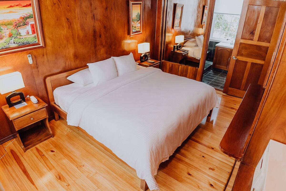 Un dormitorio con piso de madera, una cama grande, dos mesitas de noche con l ámparas y una puerta que da al baño.